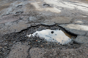 Potholes on a paved road. The road is in very poor condition. There is dirty water in the potholes.