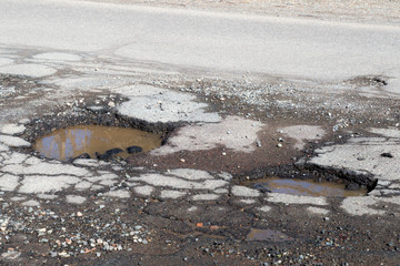 Potholes on a paved road. The road is in very poor condition. There is dirty water in the potholes.