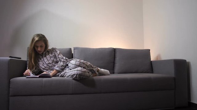 A girl lying on the couch reading a book, she is in home clothes. Spends leisure time peacefully. 4K Slow Mo