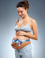 Pregnant woman with blue ribbon and touching her belly on grey background. Pregnancy, maternity, preparation and expectation concept