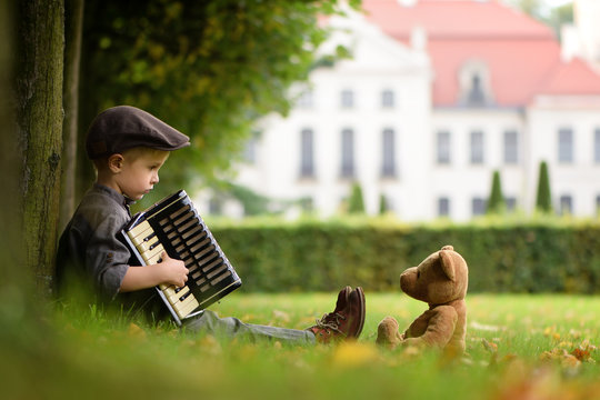 A four years old boy sitting on grass and play accordion for teddy bear 