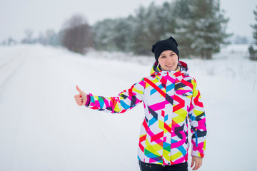 Fototapeta na wymiar Young beautiful girl in a multi-colored jacket hitchhiker catches the car on the winter road