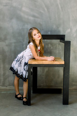 A portrait of a full-height little gir that put a hand on a chair on a gray background