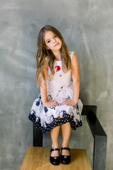 Beautiful little girl with long broun hair and blue eyes sitting on the chair on a grey background wearing a black and white spring dress