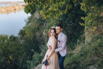 Autumn portrait of newlyweds in love, against a background of green trees, rocks and hills. Wedding photography. Stylish groom in a plaid jacket hugging a pretty bride in a beige dress with a crown.