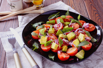Colorful and tasty salad with avocado, cherry tomatoes and mozzarella on a black dish next to a fork and knife served to eat