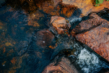 Smooth stones in spring water close-up. Clean water flow among red and orange stones. Colorful natural background of mountain spring stream with copy space. Beautiful texture of creek with wet stones.