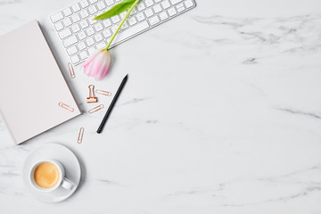 Office desk with computer, pink tulip, coffee cup and pink diary on white marble background. Flat lay. Copy space for text. Top view.