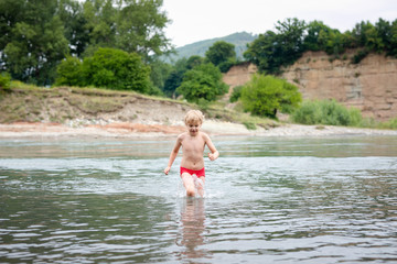 Blonde smiling boy in red swimsuit running in the mountain river