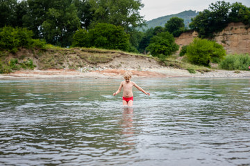 Blonde smiling boy in red swimsuit running in the mountain river