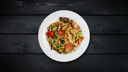 Rotating udon stir fry noodles with seafood and vegetables in a white plate on black wooden background. Top view