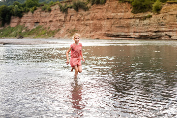 Blonde smiling girl in the pink dress playing on the side of mountain river