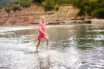 Blonde smiling girl in the pink dress playing on the side of mountain river