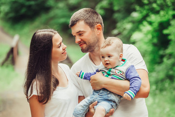 Happy loving young family with infant baby walking together in green summer park hugging each other. Family, togetherness, childhood concept