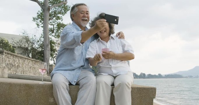 Romantic asian senior couple watching sunset and enjoying a glass of wine on beautiful tropical beach in slow motion.