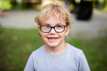 Blonde smiling boy with strabismus wearing glasses with special lens in warm park - obrazy, fototapety, plakaty