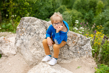 Nice funny blonde boy with strabismus wearing glasses with special lens sits on the stone in the mountains