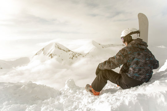 Snowboarder sit with snowboard in hands sits on big rock on mountains backdrop. Bansko, Bulgaria