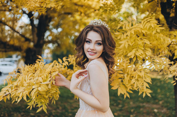 A smiling bride in a beige dress is standing in a park with yellow leaves. The princess with a...