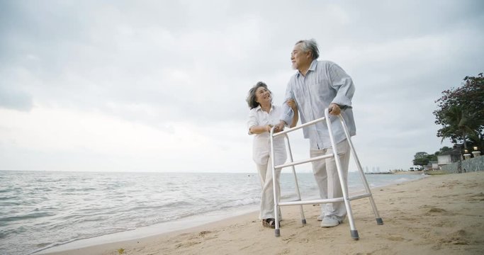Senior man exercise with walker and his wife assist him walking along beach in slow motion. People with healthcare, medical and retirement concept.