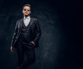 A stylish man dressed in an elegant suit with sunglasses posing with a hand in a pocket against a dark textured wall