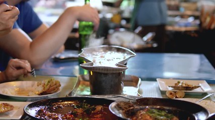 Close up shot across table of people eating steaming variety of foods in bowls & sharing out onto plates. Bokeh background of people passing in activities. - Powered by Adobe
