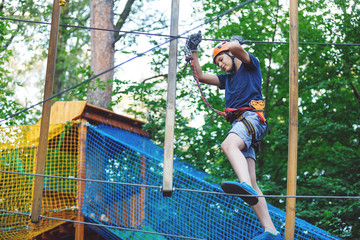 Cheerful cute young boy in blue t shirt and orange helmet in adventure rope park at sunny summer day. Active lifestyle, sport, holidays for children