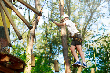 Naklejka na ściany i meble Cheerful cute young boy in white t shirt and white helmet in adventure rope park at sunny summer day. Active lifestyle, sport, holidays for children