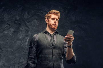 Portrait of a redhead man in formal wear using a smartphone in studio against a dark textured wall