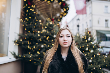 Portrait of young pretty blondie girl in christmas street decoration outdoors in coat in old town