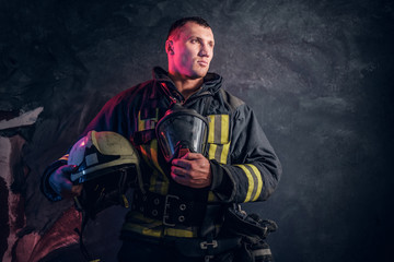 Portrait of a fireman in uniform holding a helmet and oxygen mask standing in the studio against a dark textured wall