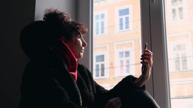 Young, modern,  middle eastern woman with curly black hair and a red sweater, relaxing on a window board, playing on her smartphone.
