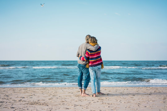 Mature couple standing on the beach with arms around, looking at the sea