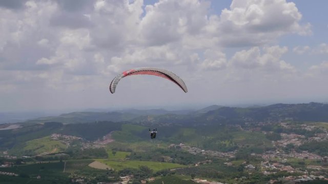 View across lush green landscape view of para glider launched from hillside floating across cloudy skies.