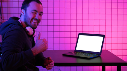 A young cheerful gamer smiling to the camera and looking at laptop. White Display.