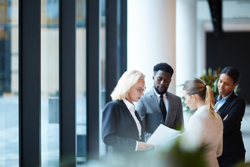 Group of elegant delegates or brokers in formalwear looking through financial papers in lounge of...