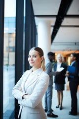 Pretty young businesswoman in white formalwear standing by window in front of camera in airport lounge