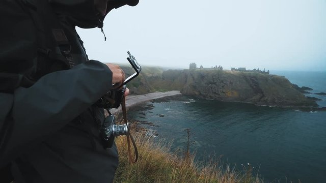 On an overcast and windy day, a man removes a roll of film from a twin lens reflex style camera, on the edge of a cliff
