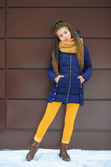 Portet of a young pretty white girl with a yellow knitted scarf in a blue jacket on the street opposite the modern brown background in winter.