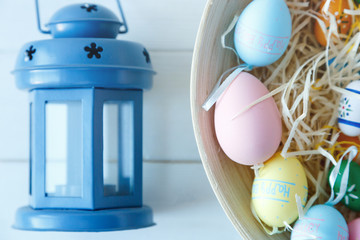 Happy Easter! Bowl with hand painted colorful eggs and blue lantern on white wooden table. Close up. Decoration for Easter, festive background.