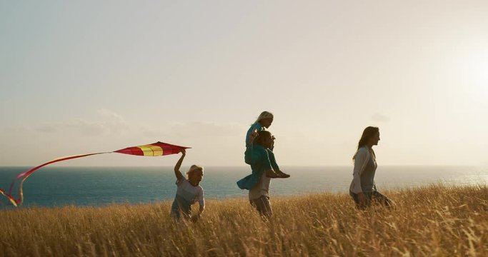 Happy family running across golden field flying kite at sunset near the ocean