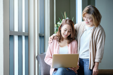 Mother hugging and teasing with her daughter while working on laptop at home, happy family concept.