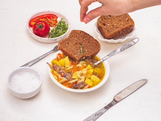 The girl's hand has a cress slice of bread. In a deep plate of freshly made soup of turkey, pasta and vegetables. Cutlery and sliced bread.