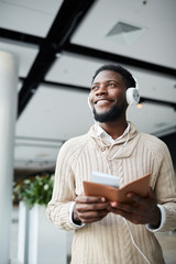 Happy African-american guy with passport and tickets listening to music in headphones during travel