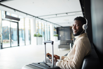 Smiling positive African-american businessman in casualwear sitting in airport lounge and enjoying...