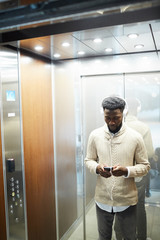 Young man in casualwear standing inside elevator and scrolling in smartphone while listening to...