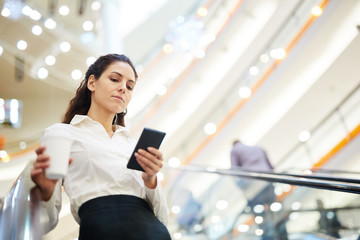 Young elegant businesswoman with drink scrolling in smartphone while moving on escalator