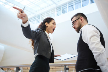 Young angry businesswoman with document shouting at confused man and pointing aside