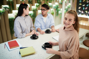 Happy high school student looking at you while having drink in college cafe on background of two classmates