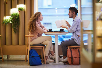 Two teenage groupmates or dates sitting in college cafe before lesson and discussing notes in copybook